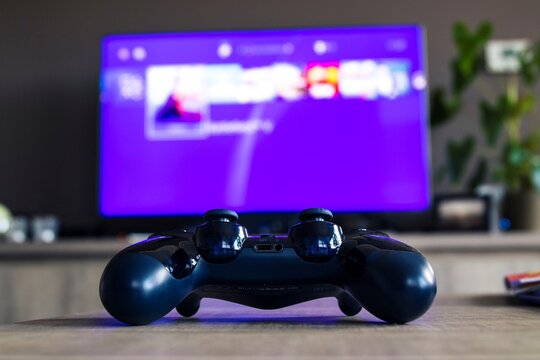 A portrait from behind a sony playstation 4 controller in front of a television with the playstation home menu displayed on it. The PS4 video game controller is turned on and ready to be played with.