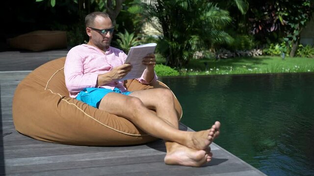 Young man reading news magazine and relaxing by the swimming pool