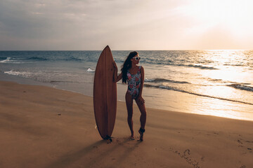 Fototapeta na wymiar Adorable attractive adult lady in bikini and sunglasses posing and standing next to a surfboard against the backdrop of the waves of the sea and sky. Summertime concept
