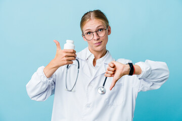 Young russian doctor woman holding pills bottle on blue showing thumbs up and thumbs down, difficult choose concept