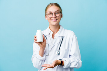 Young russian doctor woman holding pills bottle on blue holding something with both hands, product presentation.