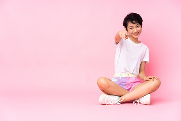 Young Vietnamese woman with short hair sitting on the floor over isolated pink background points finger at you with a confident expression