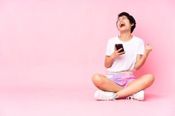 Young Vietnamese woman with short hair sitting on the floor over isolated pink background with phone in victory position