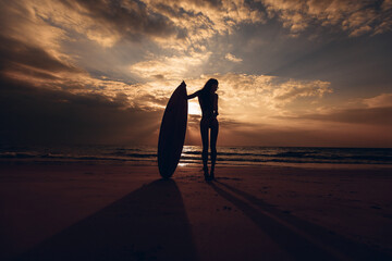 Silhouette of a slender long-legged model posing next to a surfboard on the beach against the...