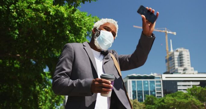 African american senior man wearing face mask holding smartphone and coffee cup hailing taxi on the 