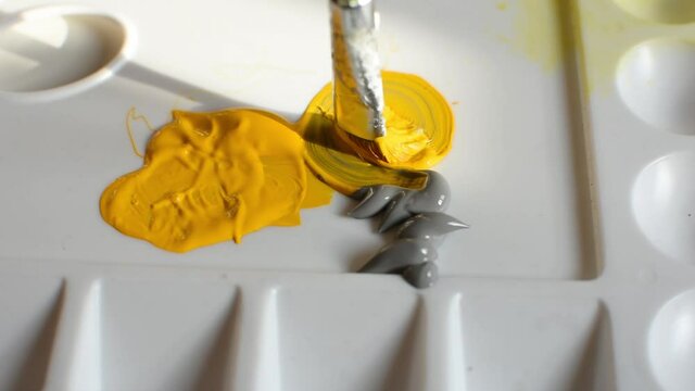 Yellow and gray acrylic paints on the palette. The artist stirs the paint with a brush with a slow movement of his hand. The creative process. Colors of 2021 ultimate gray and illuminating color