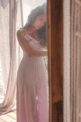 close up young girl in pink dress is standing near white panoramic window in the boho style living room at morning in sun lights, lifestyle concept