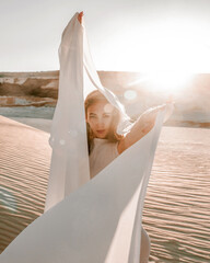 young woman is standing in gymnastic pose on the sand career in desert with white transparent cloth in hands at sunset sky background, lifestyle concept, free space