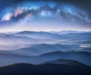 Milky Way arch over the mountains in fog at starry night in summer. Landscape with blue sky with stars, arched Milky Way, trees on the foggy hills, mountain peaks. Space and galaxy. Aerial view  