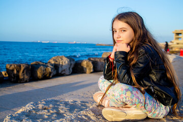 beautiful happy teenage girl with long hair in a black leather jacket and light jeans sits on the seashore on a sunny day