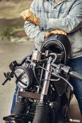 Fototapeta na wymiar Master courageous motorcycle driver in denim clothes and gloves holding a black helmet and sitting on a motorcycle. Motorcycles to order. Sports concept.