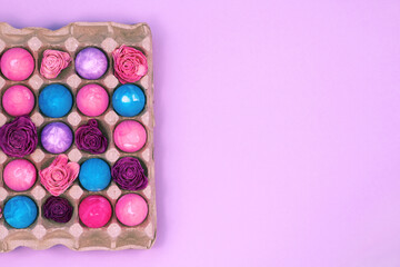 Easter eggs painted with gouache and dried roses in egg box on violet pink background, top view copy space