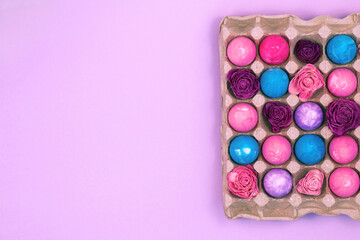 Easter eggs painted with gouache and dried flowers in egg box on violet pink background, top view copy space