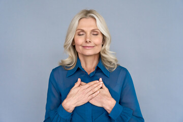 Happy mindful thankful middle aged old woman holding hands on chest meditating with eyes closed isolated on grey background feeling no stress, gratitude, mental health balance, peace of mind concept. - obrazy, fototapety, plakaty