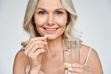 Smiling happy healthy middle aged 50s woman holding glass of water taking dietary supplement vitamin pink pill isolated on white background. Old women multivitamins antioxidants for anti age beauty. - Powered by Adobe