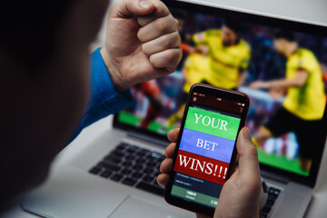 Man watching football online broadcast on his laptop and celebrate victory in betting at bookmaker's website. Focus on smartphone with message.