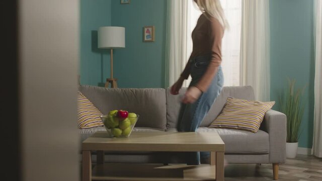 The girl in jeans, a brown sweater enters the room, takes a mobile phone and a red apple from the table, sits on the couch