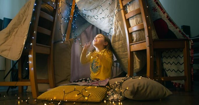 A cheerful joyful preschool girl playfully indulges and plays in a tent with Christmas lanterns sitting in the living room in the evening.