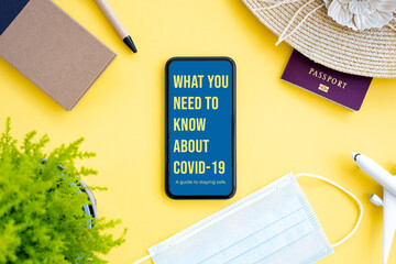 Flat lay, an aircraft model, sun hat, protective face mask, passport and a smartphone with ‘What you need to know about Covid-19, a guide to staying safe’ note, are arranged on a yellow background.