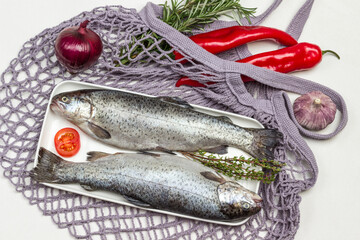 Two raw fish trout on white plate. Red pepper and sprigs of rosemary on mesh bag