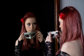 Attractive redhead young woman is looking at herself in the mirror holding a carnival mask. Horizontally. 