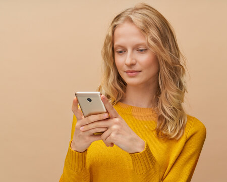 Pretty female looking social media on mobile phone standing near beige background. Natural realistic beauty, fresh face of youthful woman. Portrait of beautiful blonde in yellow jumper with cell phone