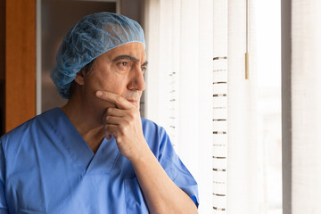 Doctor on a coffee break. Mature male surgeon thinking during a break. He has a pensive gesture looking out the window. Wearing a surgical cap.