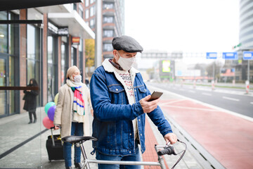 Senior man with bicycle and smartphone outdoors on bus stop in city, coronavirus concept.