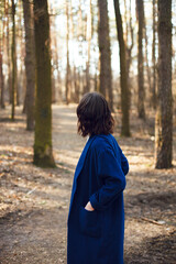 a peron in a blue long coat stands sideways and looks into the distance in a pine forest