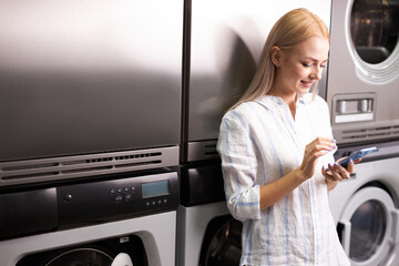 Laundry, smiling woman holding phone, washing clothes via app. Young caucasian female is ready to wash clothes