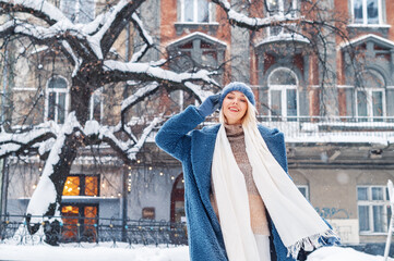 Winter, Christmas, New Year holidays, travel concept: happy smiling woman posing at street of European city. Snowfall. Model wearing blue faux fur coat,  hat, white scarf. Copy, empty space for text