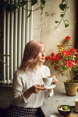 young woman in a white blouse dreamily looks to the side and holds a white cup with coffee in a cafe restaurant