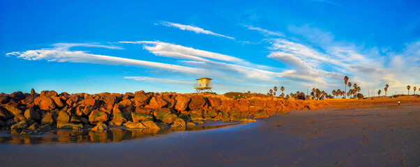 Cloud streaked-sky on a beach with a breakwater and lifeguard tower, birds flying through the air in a relaxing evening sea shore scene.  Golden rocks, white clouds and birds are in the sky.