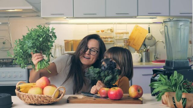 Funny young mom and child daughter making bunches fresh herbs having fun. Happy kid helping mum in kitchen laughing, play, dance. Cheerful vegan family enjoying cooking activity