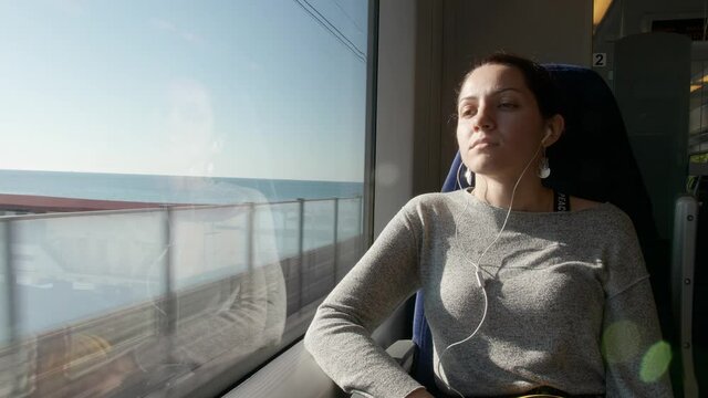 a young girl sits on a train traveling by the sea, at the window in white monitors, illuminated by the hot summer sun. outside the train window you can see the big blue ocean on a clear summer day