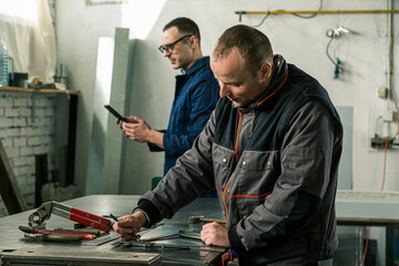 A tinsmith is measuring in his workshop with a colleague engineer who is taking notes on tablet