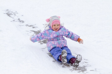 A little pretty girl in a purple overalls slides down an ice slide, winter children's games, a...