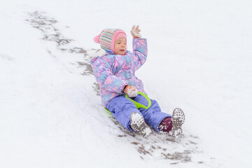A little pretty girl in a purple overalls slides down an ice slide, winter children's games, a...