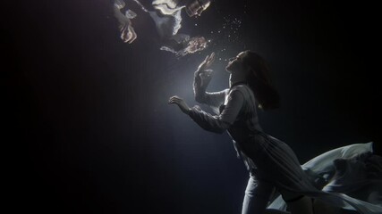 A beautiful woman in a long flowing dress floats under the water on a dark background, she swims to the surface of the water to her reflection, creating a lot of bubbles.