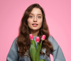 Young woman with tulips on a pink background. Blue eyes, long hair and surprise on her face.