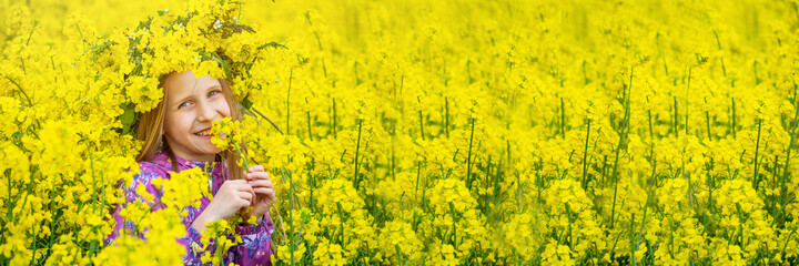 Happy girl with long light hair and grey eyes is standing in the field of yellow flowers. The wreath from canola is beautify the head. Rural landscape of blooming rapeseed.