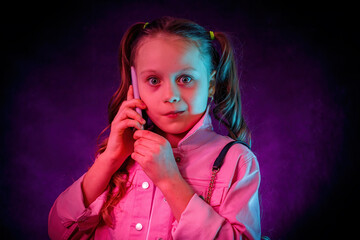 The little girl is talking on the phone. A surprised, startled face. Studio, neon light, gel filters, turquoise, magenta, purple.