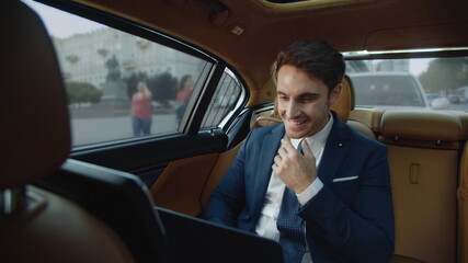 Smiling business man chatting on laptop computer in luxury automobile.