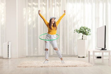 Young woman spinning a hula hoop at home in a living room - obrazy, fototapety, plakaty