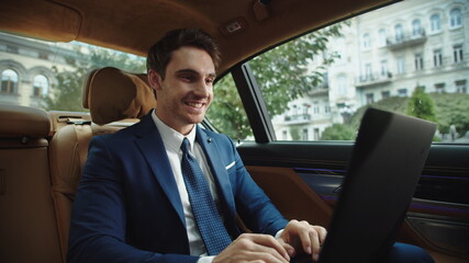 Cheerful businessman typing laptop in luxury car. Male ceo celebrating success