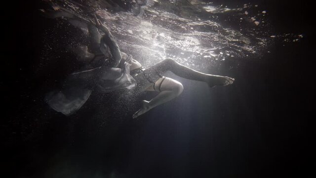 A beautiful woman in a long dress hovers on her back under the water on a dark background, she swims to the surface of the water to her reflection, creating a lot of bubbles.
