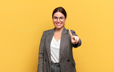 young pretty woman smiling and looking friendly, showing number one or first with hand forward,...