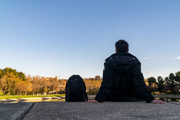 Traveler man in a black coat and backpack, sitting in front of a pond while resting.