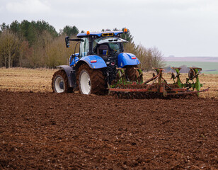 New Holland tractor cultivates a dark red mud field in winter