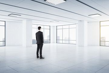 Businessman in black suit in empty spacious hall room with light walls, big windows